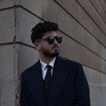 A fashionable bearded man poses stylishly in a suit and sunglasses, leaning against an outdoor wall.