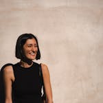 A joyful woman in a black outfit smiles by a sunlit beige wall.