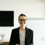 Caucasian woman in black blazer with eyeglasses, smiling in classroom.