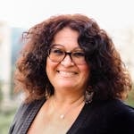 Portrait of a woman with curly hair and glasses smiling warmly in an outdoor setting.